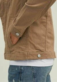 Brun canvasjacka med en knäppt manschett, sidofickor och en enkel nederkant. Bärs över en vit skjorta och denimjeans.