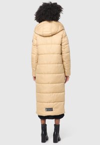 Long, beige puffer coat with a hood, featuring horizontal stitching, a knee-length design, and a label at the hem. Worn with black ankle boots.