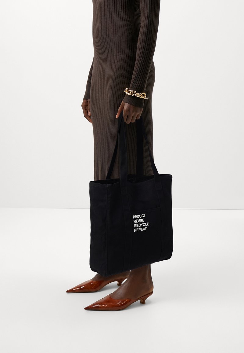 Czarna torba na zakupy z tekstem "REDUCE REUSE RECYCLE REPEAT" na białym tle. Długie uchwyty, prostokątny kształt, trzymana przez osobę w długiej brązowej sukience.
