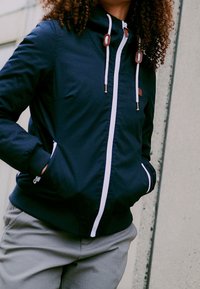 Granatowa kurtka wiatrówka z białymi zamkami błyskawicznymi i sznurkiem do ściągania, wyposażona w dwie przednie kieszenie i wykończenie z teksturowanej tkaniny.