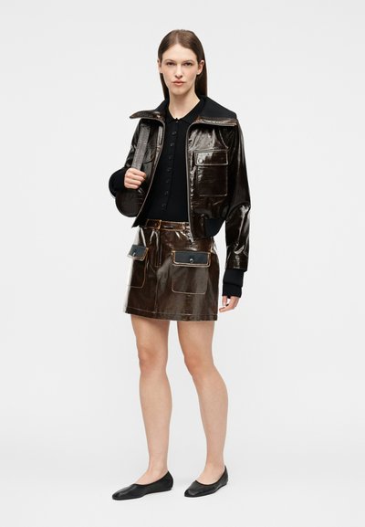 Femme portant une veste en cuir brillant marron foncé et une jupe assortie avec des poches avant, un haut noir et des ballerines noires, debout devant un fond blanc.