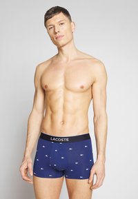 Temno modri boksericii z belimi vzorci logotipa Lacoste, s črnim elastičnim pasom z besedilom "LACOSTE". Narejeni iz mehke bombažne tkanine.