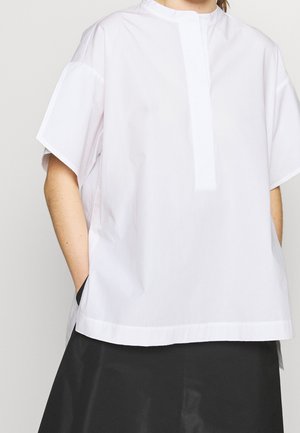 Hvid oversized skjorte med korte ærmer og skjult frontlukning, båret udenfor en sort nederdel, model med hænderne i lommerne.