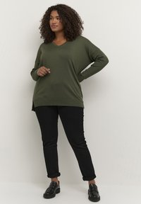 Maglione verde oliva a V con maniche lunghe e spacchi laterali, abbinato a pantaloni neri con risvolto e mocassini neri. Tessuto liscio, vestibilità morbida.