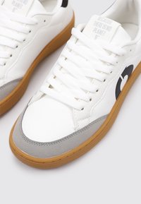 Baskets blanches en cuir avec un capuchon en suède gris, logo noir sur le côté et semelle en caoutchouc beige. Comprend six œillets pour lacets et lacets plats blancs.