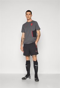 Grå atletisk skjorte med røde detaljer, sort panel, parret med sorte shorts. Har tatoveringer, sorte sokker og sorte sportsko.