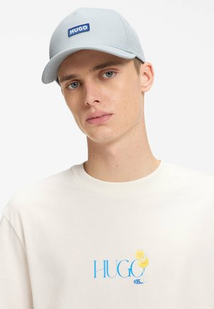 Jeune homme portant une casquette HUGO gris clair et une chemise blanche avec un logo HUGO bleu et un dessin de boisson jaune sur la poitrine.