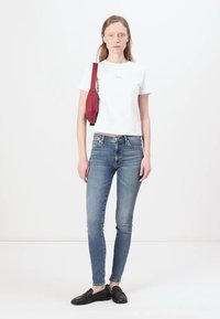 Tricou alb scurt și blugi skinny albaștri. Geantă roșie, tip crossbody, cu un design elegant. Pantofi negri, fără toc. Outfit simplu și casual. Fundal minim.