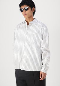 Weiß gestreifte Button-Down-Bluse in lockerer Passform, mit Taschen auf der Brust, klassischem Kragen und langen Ärmeln. Getragen mit schwarzen Hosen.