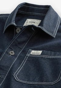 Mørk denimskjorte med spidse krave, kontrasterende hvid syning, metalknapper og en brystlomme med et mærke.