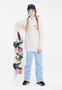 Witte snowboardjas, lichtblauwe broek, staand met een kleurrijke snowboard met een grafisch ontwerp en zwarte bindingen. Zwarte goggles op het hoofd.