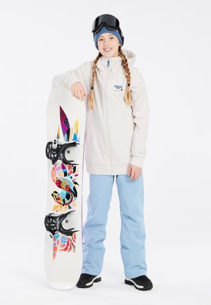 Weiße Snowboardjacke, hellblaue Hose, steht mit einem bunten Snowboard, das ein grafisches Design und schwarze Bindungen aufweist. Schwarze Skibrille auf dem Kopf.