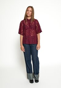 Blusa in pizzo bordeaux con maniche corte, design con bottoni sul davanti, abbinata a jeans in denim a gamba larga con risvolti in contrasto e scarpe nere.