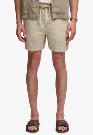 Uomo che indossa pantaloncini beige con coulisse, sandali marroni a ciabatta e una camicia marrone chiaro con maniche rimboccate, mostrato dalla vita ai piedi.