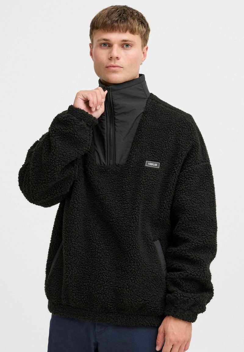 Black fleece pullover with a high collar and half-zip, featuring a smooth panel at the neck and logo patch on the chest.