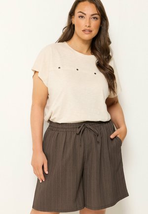 Kvinde iført en beige kortærmet top med metalnitter og løse brune shorts med snører, poserende med den ene hånd i lommen.