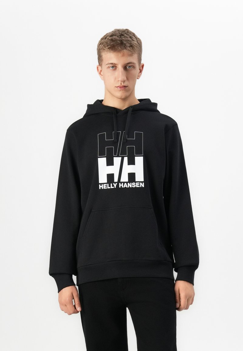 Giovane uomo che indossa un hoodie nero Helly Hansen con logo bianco, in piedi contro uno sfondo bianco uniforme, rivolto in avanti.