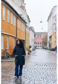 Manteau noir long avec un col montant, doté de manches amples et de poches latérales, associé à un jean bleu large et des chaussures sportives blanches et noires.