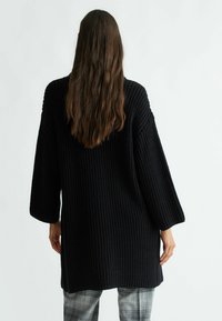 Černý pletený svetr s volným střihem, dlouhými rukávy, ribovanou texturou a zakřiveným lemem, spárovaný s kostkovanými kalhotami.