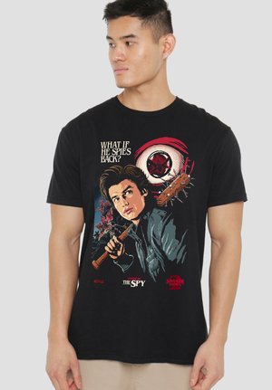 Young man wearing black T-shirt with graphic of a boy holding spiked bat and text "What if he spies back?" from Stranger Things.