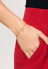 Złocony bransoletka łańcuchowa z motywem serca i symbolu nieskończoności. Bransoletka jest noszona na dłoni spoczywającej w kieszeni czerwonych spodni.