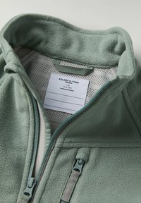 Fleece-takki vaaleanvihreänä, jossa on korkea kaulus, täysipitkä vetoketju, verkkomateriaali vuorissa ja tekstiilien hoito-ohjeet näkyvästi sisäpuolella.