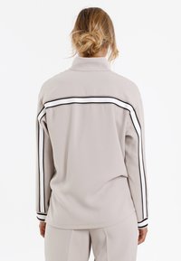 Hellegrauer Pullover mit hohem Kragen, ausgestattet mit schwarz-weißen gestreiften Akzenten über den Rücken und die Ärmel. Glattes, strukturiertes Material.