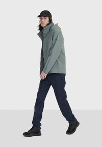 Veste imperméable vert clair avec capuche, poignets à boutons et poches latérales, associée à un jean en denim foncé et des baskets noires.