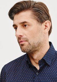 Granatowa koszula z guzikami, ozdobiona małym, powtarzającym się wzorem w jasnobrązowym kolorze, z wyraźnym kołnierzykiem i teksturowanym materiałem.