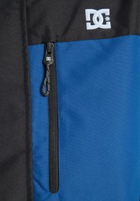 Niebieska i czarna kurtka z zamkiem błyskawicznym, teksturowany materiał oraz wyraźne białe logo na klatce piersiowej.