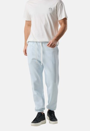 Homme portant un t-shirt blanc à motif, un pantalon de jogging bleu clair avec cordon de serrage et des baskets à plateforme noires, debout devant un fond uni.