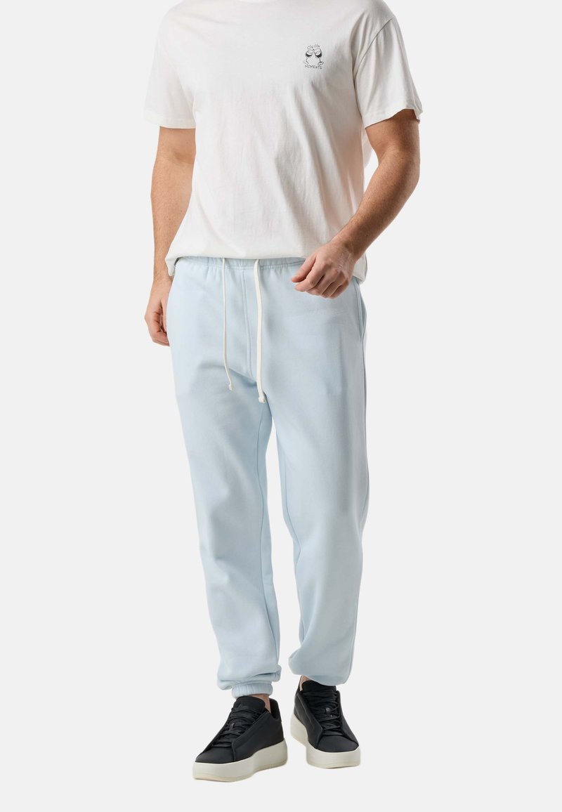 Άνδρας που φοράει λευκό μπλουζάκι με γραφικό σχέδιο, ανοιχτό μπλε παντελόνι φόρμας με κορδόνι και μαύρα πλατφορμ παπούτσια, στέκεται μπροστά σε απλό φόντο.