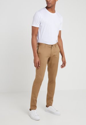 Mężczyzna ubrany w prosty biały t-shirt z krótkim rękawem, beżowe spodnie typu slim fit i białe sneakersy, stoi na neutralnym tle.