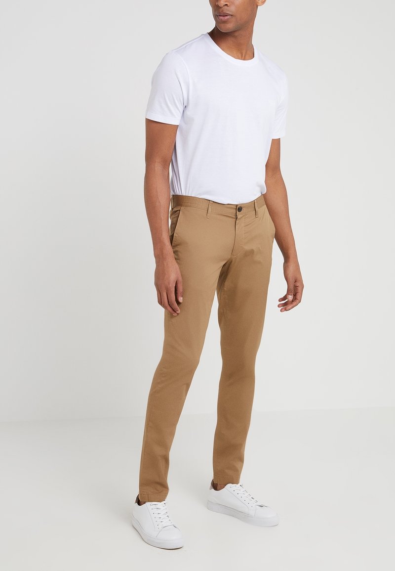 Homme portant un t-shirt blanc uni à manches courtes, un pantalon beige slim et des baskets blanches, debout devant un fond neutre.