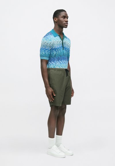 Modrá a zelená polokošile s krátkými rukávy, spojená s olivovými zelenými kraťasy. Bílá teniska a ponožky doplňují vzhled.