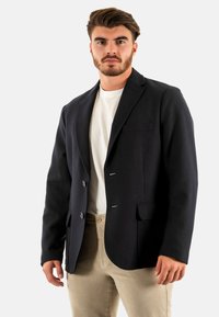 Blazer blu navy con due bottoni, alette affilate, tasca sul petto e tasche laterali. Indossato sopra una t-shirt bianca e pantaloni beige. Texture del tessuto morbida.