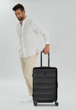 Homme en chemise blanche et pantalon beige tenant une valise noire à roulettes avec poignée dépliée sur fond gris.