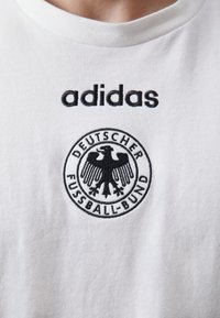 Balta medvilninė marškinėliai su juodu "adidas" logotipu ir apvaliu išsiuvinėtu emblemą, kuriame pavaizduotas Vokietijos futbolo sąjunga ir erelis.
