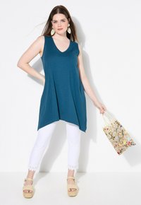 Tunica senza maniche color teal con scollatura a V, abbinata a pantaloni bianchi a cropped con pizzo. Indossata con sandali con zeppa e una borsa a tracolla floreale.