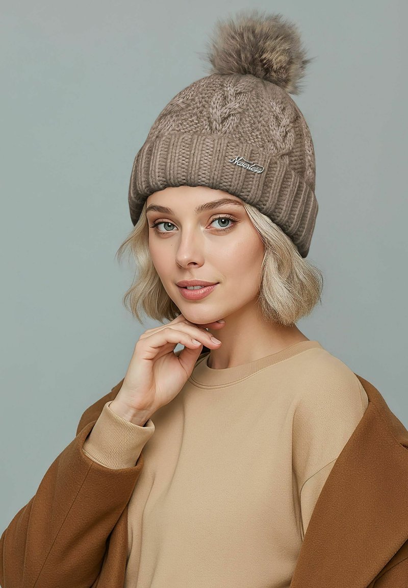 Knit beige beanie with a fluffy pom-pom on top, featuring a braided texture and a logo patch on the side. Worn with a beige top.