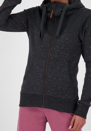 Gros plan sur une personne portant un sweat à capuche zippé gris foncé avec de petits motifs de cœurs roses et un pantalon rose, la main reposant le long du corps.