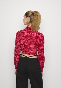 Top curto vermelho com um acabamento texturizado e padrão, mangas longas, gola alta e costas expostas com laços ajustáveis.