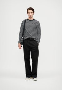 Fekete-fehér csíkos, hosszú ujjú póló, fekete nadrág, sötétkék sportcipő. A modell egy keresztpántos táskával áll, egyszínű háttér előtt.