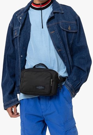 Persona che indossa una giacca di jeans blu, una camicia azzurra e pantaloni cargo blu, con una borsa a vita nera Eastpak attorno alla vita.