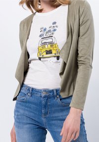 Oliven grøn letvægtsjakke over en lys cremefarvet t-shirt med et gult vintage bilgrafik og blåt blomsterdesign, parret med blå jeans.