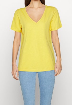 Vrouw draagt een felgeel V-hals T-shirt met korte mouwen, gecombineerd met lichtblauwe strakke jeans, poseert tegen een effen witte achtergrond.