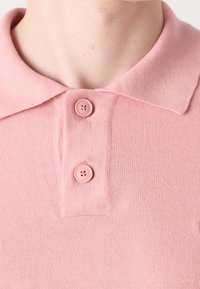Gros plan d'une personne portant un polo en maille rose avec un col et deux boutons roses fermés au niveau de l'encolure.