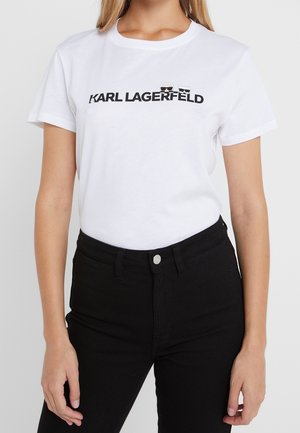 Persoon draagt een wit Karl Lagerfeld T-shirt met zwart logo en twee karakterpictogrammen boven een zwarte broek met hoge taille.