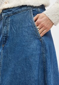 Džinsa svārki ar augstu vidukļa dizainu, zili, ar sānu kabatām un vaļīgu piegriezumu. Tekstūra ir gluda ar redzamām šuvju detaļām.