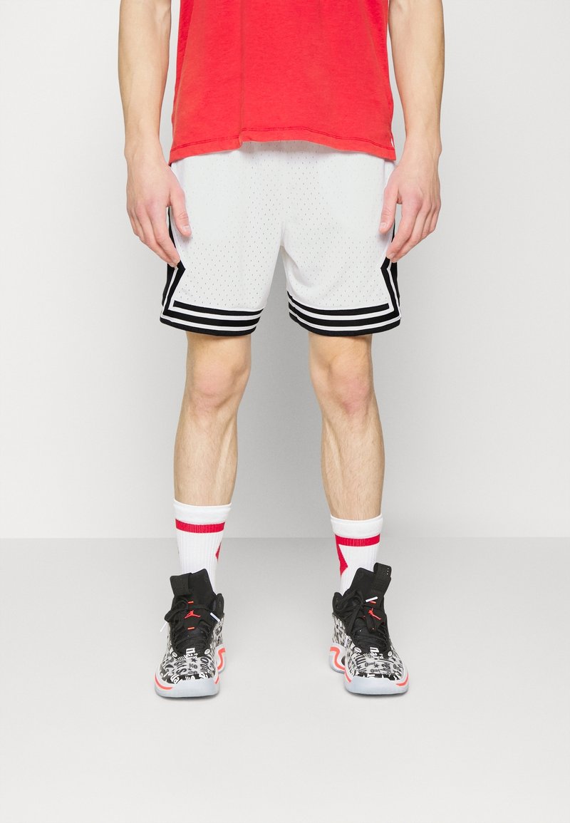 Man wearing white basketball shorts with black stripes, white socks with red patterns, black-and-white high-top sneakers, and a red shirt cropped out.
